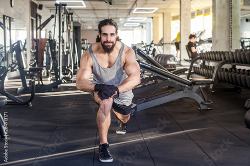 Portrait of young adult sportman athlete with long curly hair working out in gym, squating on one knee, stretching after training, doing exercises for legs, squatting. looking at camera and smiling © khosrork