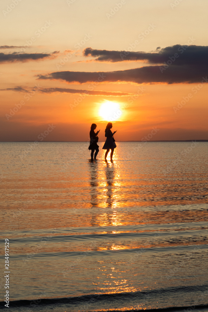silhouette of two women with cell phones in hands on the beach at sunset