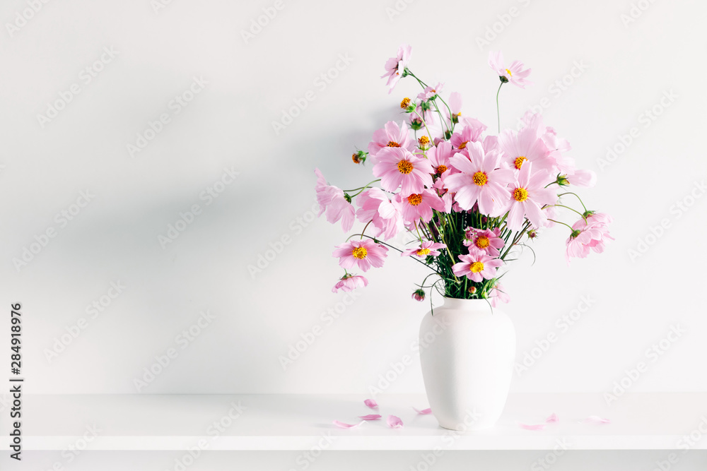 Fresh summer bouquet of pink cosmos flowers in white vase on white wood shelf on white wall background. Floral home decor. Fresh summer bouquet of pink cosmos flowers in white vase on white wood shelf on white wall background. Floral home decor.
