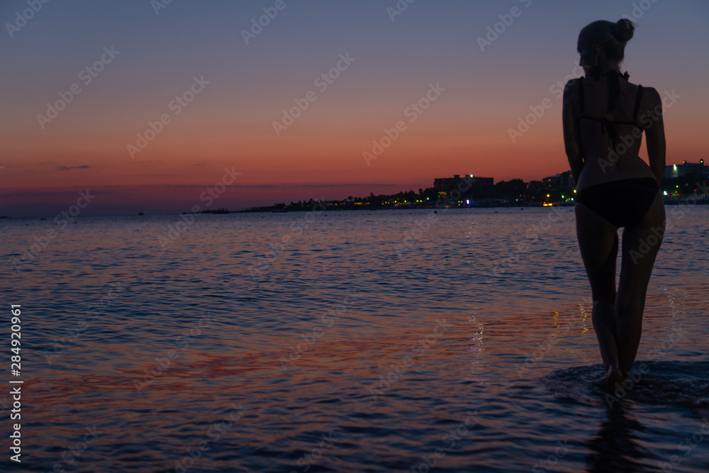 Fototapeta premium Sexy beautiful woman stands on the beach at orange sunset in the warm sea with silhouette