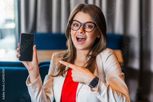 Portrait of shocked beautiful stylish young woman in glasses sitting, showing and pointing at mobile smart phone screen and looking at camera with surprised face. indoor studio shot, office background photo