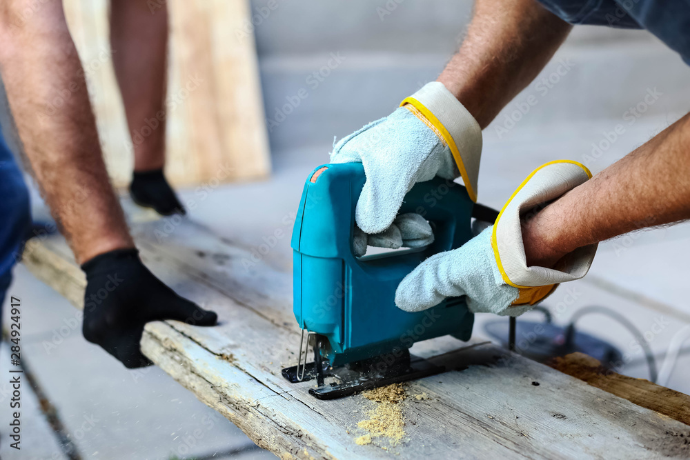  Two carpenters in working process in the carpentry workshop. Carpenter using fretsaw for cutting wooden boards. Construction details of male worker or handy man with power tools.