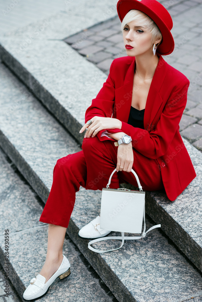 Outdoor full body fashion portrait of young elegant woman with short blonde  hair, wearing red suit, blazer, trousers, hat, white loafers, wrist watch,  holding small bag. Model posing in street Stock Photo |