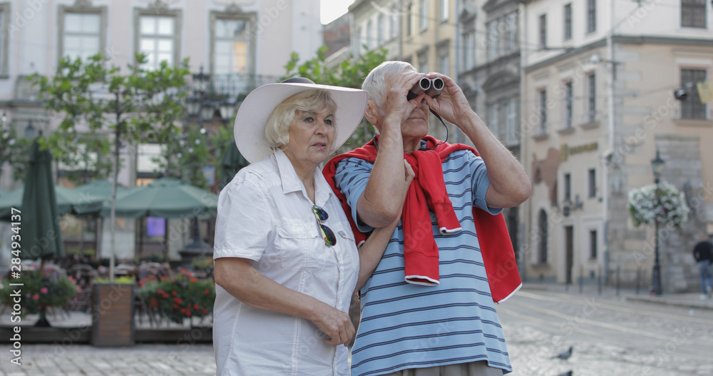 Senior male and female tourists walking in town center and looking in binoculars