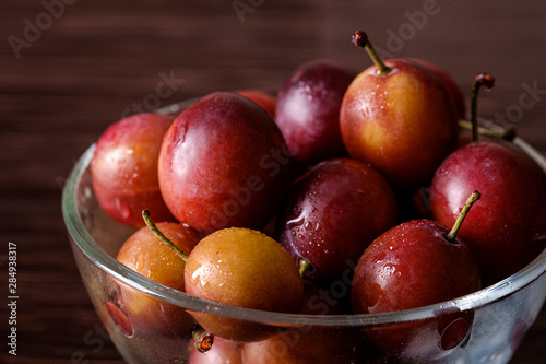 Ripe sweet plum fruits with water drops in glass bowl on dark moody wood table background