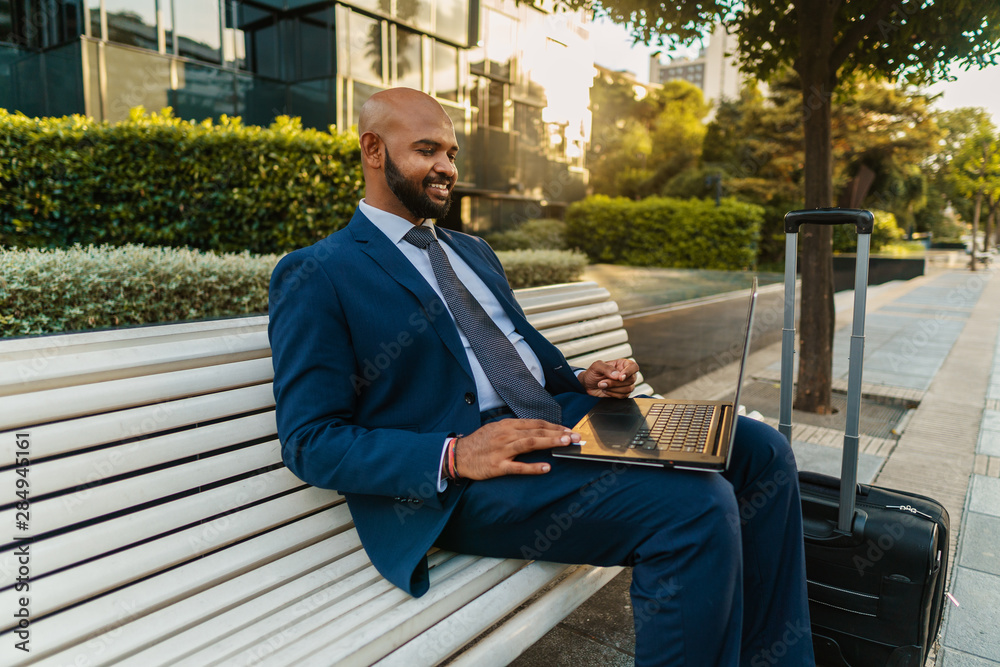 Fototapeta premium Indian businessman holding laptop notebook wearing blue suit near office. Freelance business work concept. His suitcase is near bench