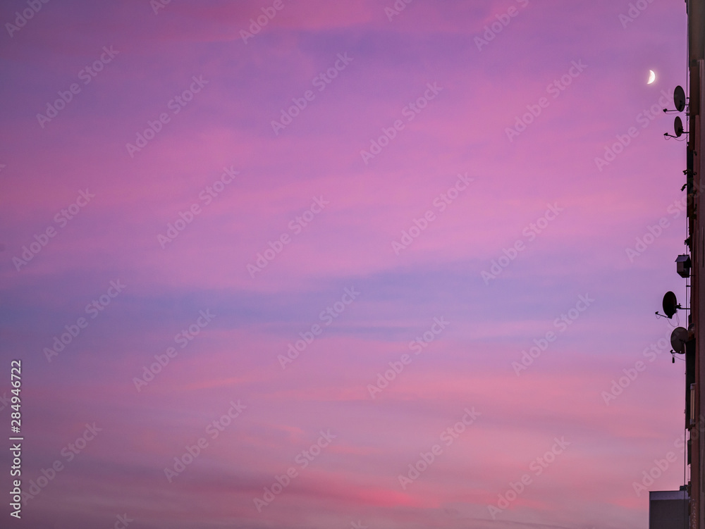 Beautiful colorful clouds at sunset with a moon in the corner of the image. Framed by the edge of the house with satellite antennas.