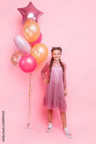 Happy and beautiful teen girl holding a bunch of colorful balloons and looking at camera with smile on pink background