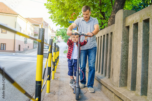 Small boy son child on the electric push kick scooter with his grandfather senior man helping him to learn to ride on the sidewalk