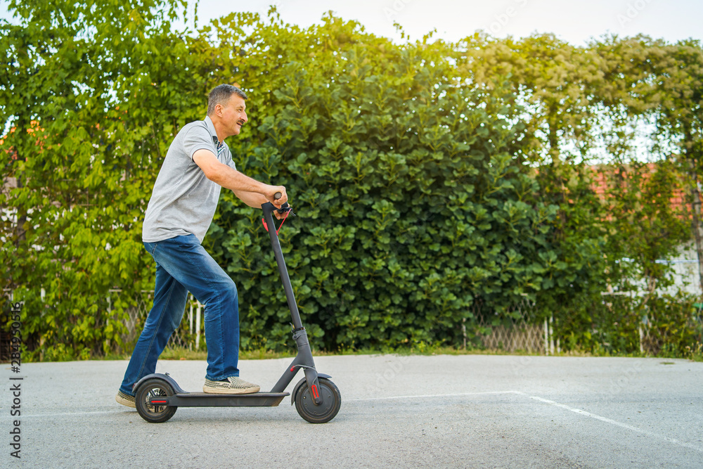 Senior man riding kick push scooter on the asphalt in front of the bushes green fence in a summer day Senior man riding kick push scooter on the asphalt in front of the bushes green fence in a summer day