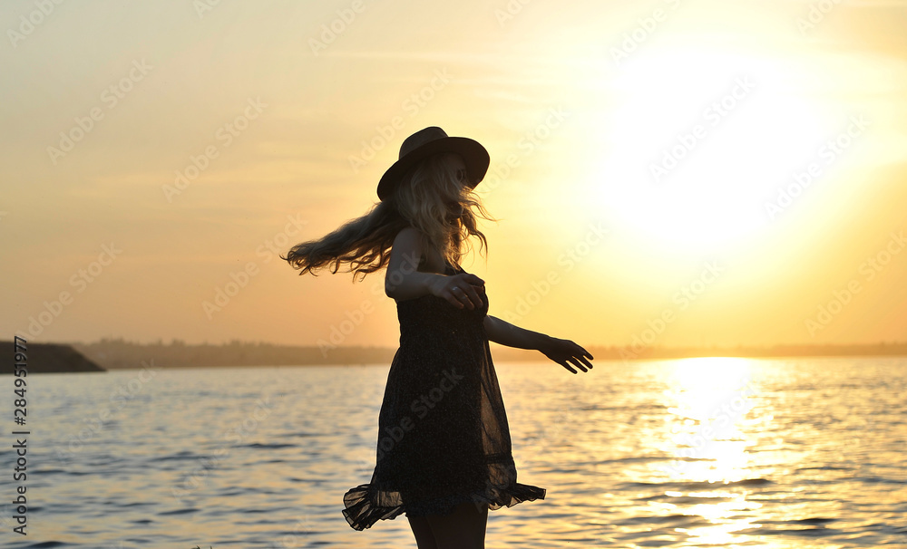 Fototapeta premium Stylish woman in hat walks on the beach at sunset.