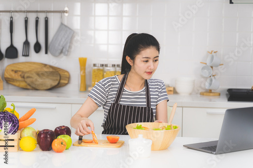 Beautiful young woman is learning to cook healthy food online by the internet from a laptop in white kitchen with many fresh vegetables and fruits on table.