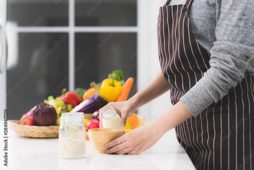 Close up woman 's hand pouring milk from bottle in wooden bowl with cereal in white kitchen. Close up woman 's hand pouring milk from bottle in wooden bowl with cereal in white kitchen.