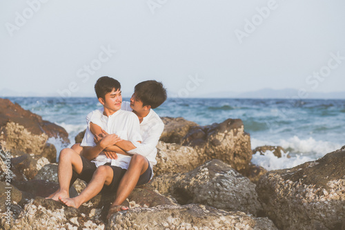 Homosexual portrait young asian couple sitting hug together on rock or stone in the beach in summer, asia gay going tourism for leisure and relax with happy in vacation at sea, LGBT legal concept. © N_studio