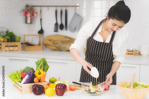 Beautiful asian woman or housewife is cooking salad for family in white kitchen with many fresh vegetables and fruits on table.