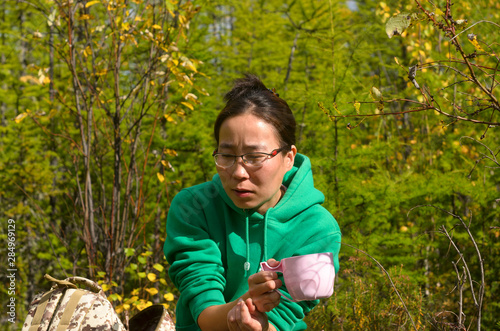 Thoughtful Yakut Asian girl tourist with glasses sitting with a Cup of tea next to a backpack in the wild forest of Yakutia.