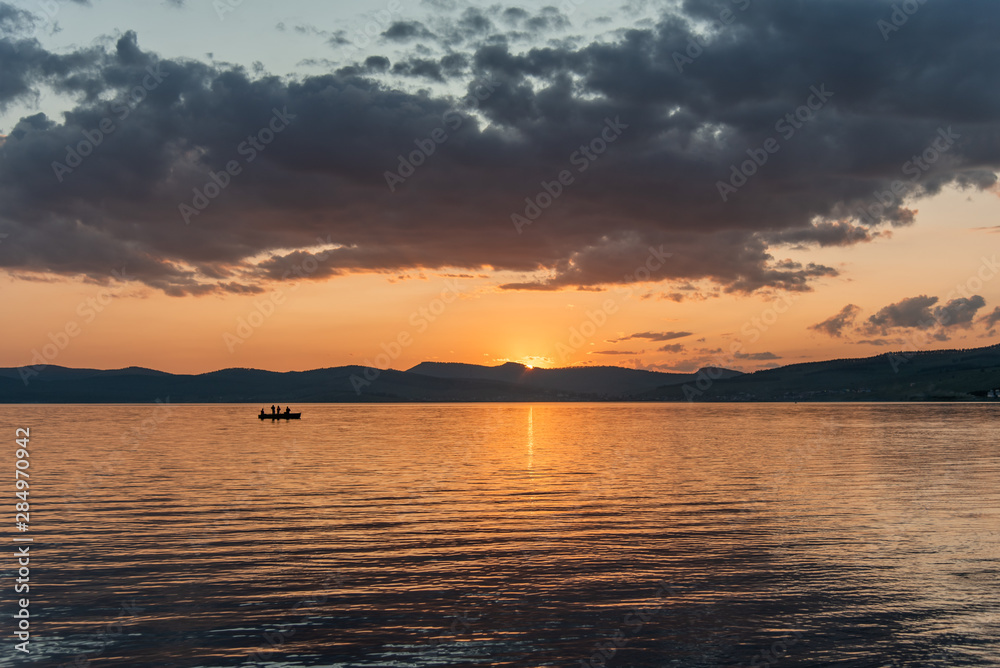 Burning red sunset on the river. Men riding boat. Silhouette of mountains on the background. Beautiful clouds in the sky. Burning red sunset on the river. Men riding boat. Silhouette of mountains on the background. Beautiful clouds in the sky.