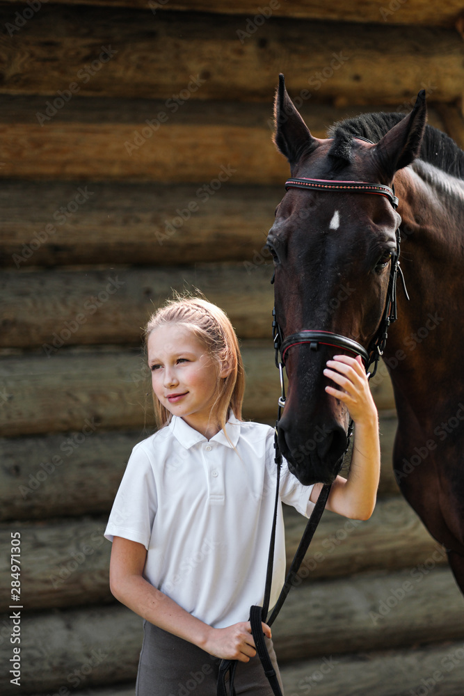 Obraz premium A teenage girl jockey stands next to a brown horse and hugs her. Against the background of a wooden wall stables.