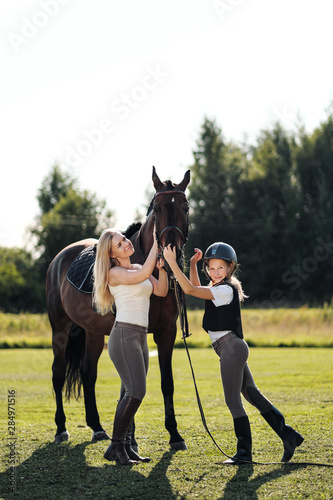 Mother and daughter riders and jockeys on a green field hug a brown horse. © spaskov