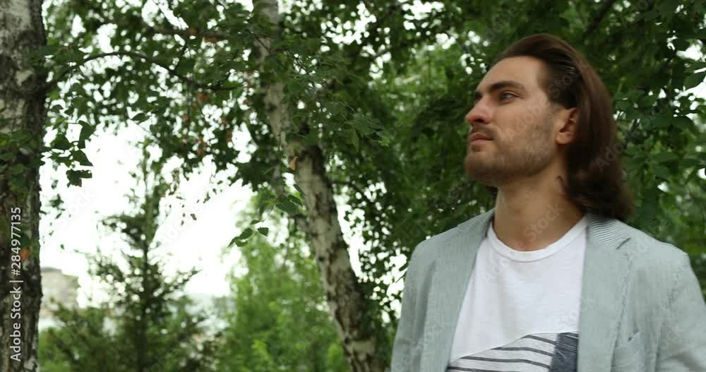 An attractive young man with a beard and mustache looks around and looks at his watch, smiles in the Park. Men's fashion, beauty. Summer day. An attractive young man with a beard and mustache looks around and looks at his watch, smiles in the Park. Men's fashion, beauty. Summer day.