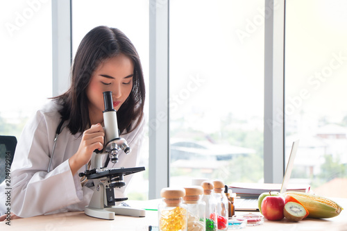 Young Asian female nutritionist doctor testing food samples for good health with microscope on wooden table in laboratory, healthy concept