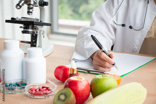 Close up photo of Young Asian female nutritionist doctor work at wooden table and writing medical records on a clipboard with fresh fruit on foreground in laboratory, healthy concept