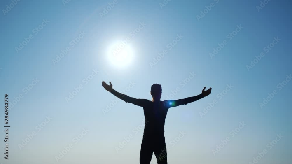 Young long-haired inspired man raises his hands up standing on the top of a mountain above the sea against beautiful blue sky. Silhouette of a happy hiker man standing on the summit. Young long-haired inspired man raises his hands up standing on the top of a mountain above the sea against beautiful blue sky. Silhouette of a happy hiker man standing on the summit.
