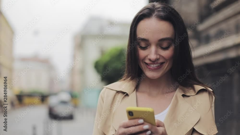 Portrait of Beautiful Young Girl Holding Mobile Phone Chatting Standing Loking into Camera and Smiling at City Background. Close up. Portrait of Beautiful Young Girl Holding Mobile Phone Chatting Standing Loking into Camera and Smiling at City Background. Close up.