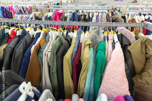 Row multi-colored of adult, male and female winter jackets hang on hanger clothes rack in retail fashion store or second hand outlet shop.