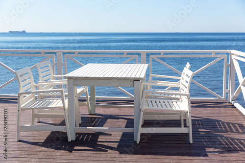 Four white, wooden chairs and a table stand on the wooden flooring in the cafe on the veranda near the blue sea in Sunny weather in summer.