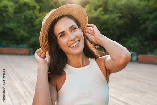 Image of joyful middle-aged woman smiling and looking at camera while walking in summer park