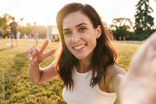 Image of happy middle-aged woman showing peace sign and taking selfie photo in summer park photo