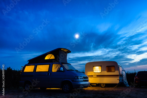 Принти на полотні A camper van with its pop top roof open parked next to a vintage caravan in a campsite at nightfall, both illuminated from inside and the moon glowing above in a stormy sky
