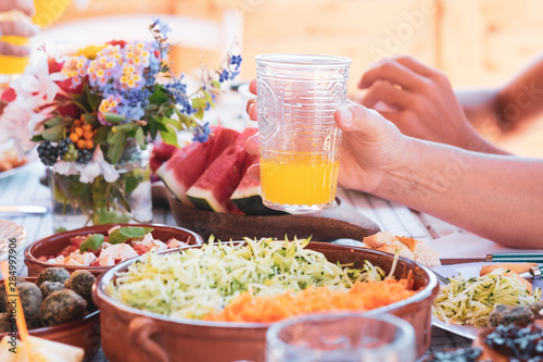 Close-up of wooden table rich of vegetables and watermelon. Caucasian arms with drinking glass. Brunch outdoor for the family