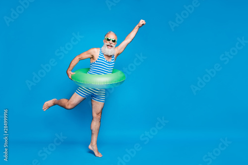 Fototapeta Naklejka Na Ścianę i Meble -  Full length photo of excited retired pensioner with grey hair raising his fist holding toy circle wearing striped swimwear glassses isolated over blue background
