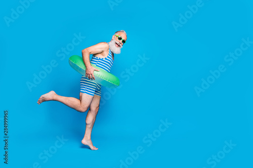 Full length photo of cheerful retired person with eyeglasses eyewear white hair holding toy ring wearing striped bathing suit isolated over blue background