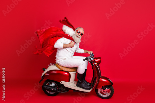 Full length profile side photo of funny pensioner carrying bag with gifts sitting on motorcycle hurrying wearing white sweater pants boots isolated over red background