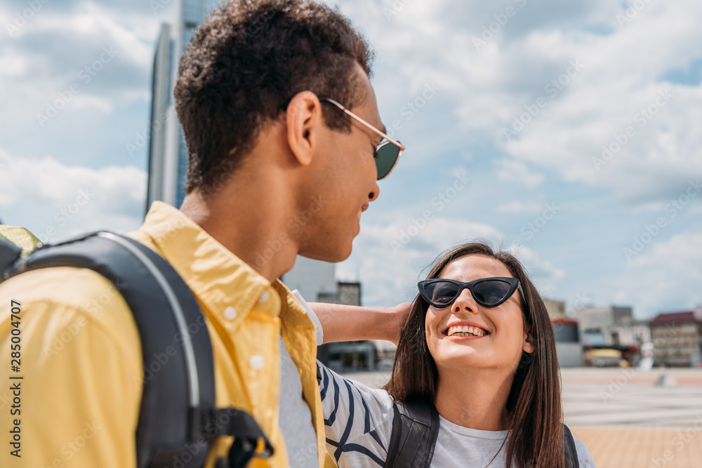 Woman in sunglasses smiling and looking at bi-racial friend with backpack