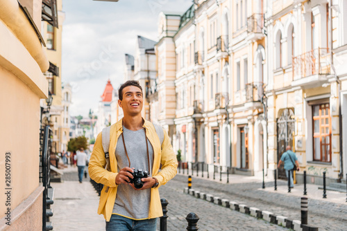 happy mixed race man smiling while holding digital camera near buildings