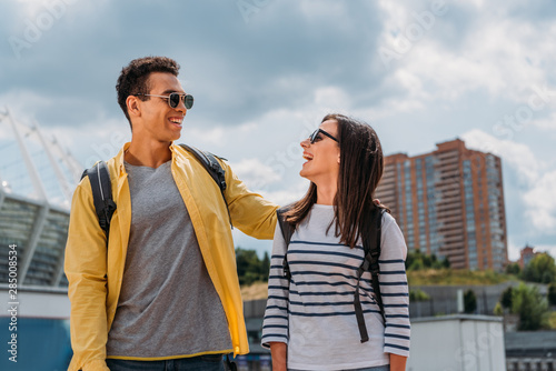 Cheerful interracial friends in sunglasses looking at each other and smiling with backpacks