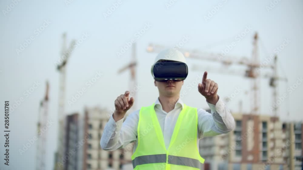 A man in virtual reality glasses helmet and vest on the background of construction controls the hands of the interface and checks the quality of construction and development A man in virtual reality glasses helmet and vest on the background of construction controls the hands of the interface and checks the quality of construction and development