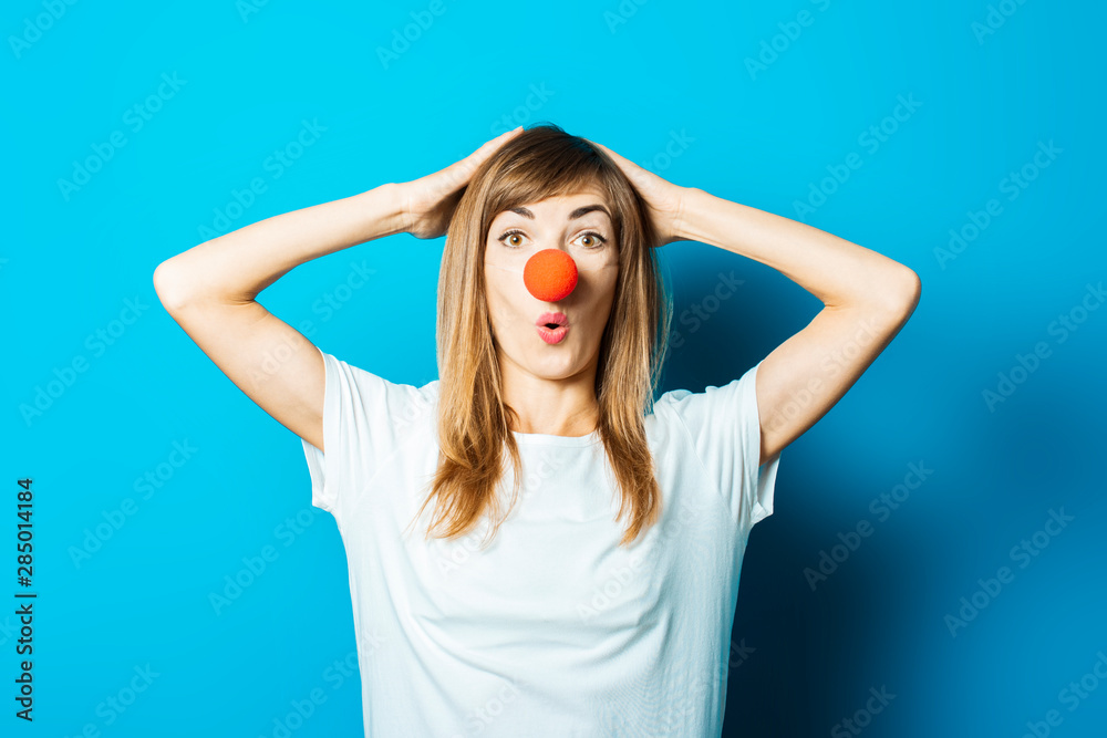 Beautiful young woman in a white T-shirt and red nose of a clown with a surprised face on a blue background. Concept party, costume, red nose day Beautiful young woman in a white T-shirt and red nose of a clown with a surprised face on a blue background. Concept party, costume, red nose day