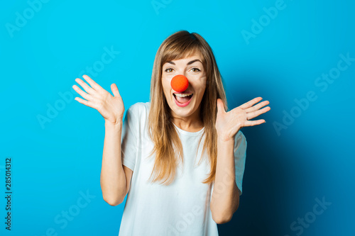 Beautiful young woman in a white T-shirt and a red clown nose smiles and makes hand gestures on a blue background. Concept party, costume, red nose day