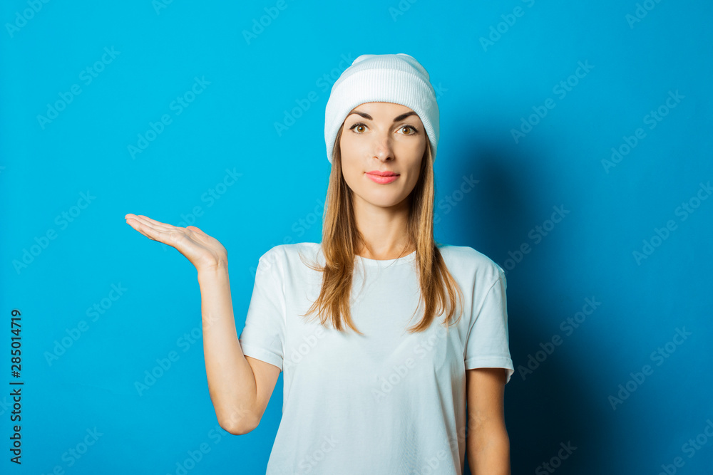 Young woman in white hat and t-shirt makes a hand gesture on a blue background Young woman in white hat and t-shirt makes a hand gesture on a blue background