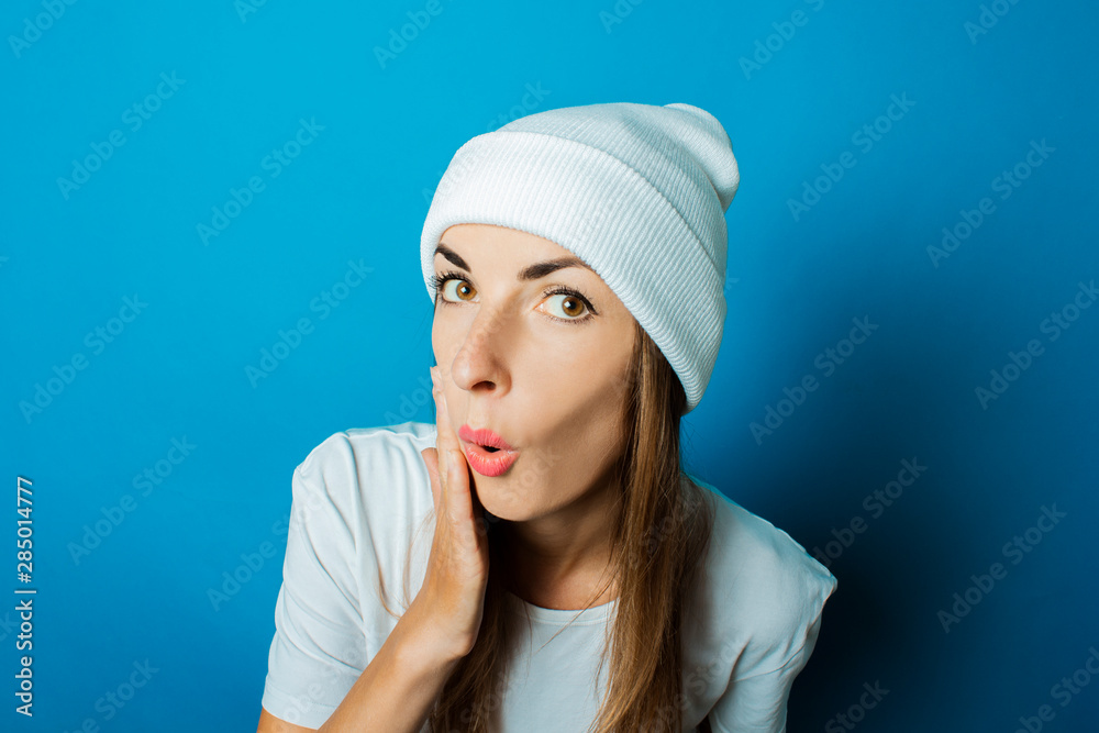 Young woman in a white hat and a T-shirt with a surprised face on a blue background Young woman in a white hat and a T-shirt with a surprised face on a blue background