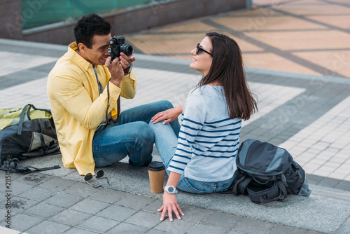 Bi-racial man sitting on stairs and taking photo of woman in sunglasses with backpack
