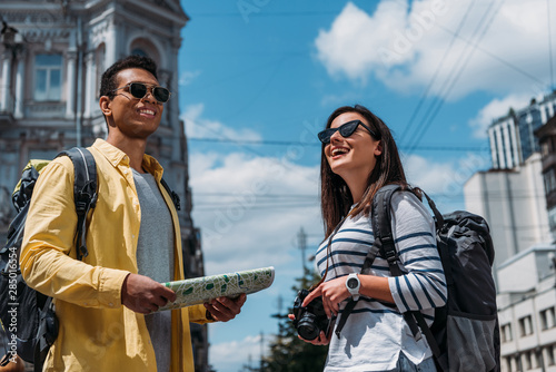 Woman in sunglasses with digital camera near bi-racial friend with map and backpack