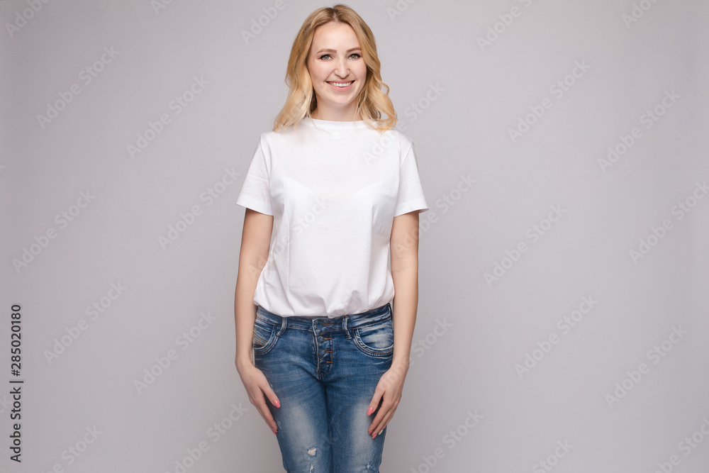 View from front of beautiful slim woman wearing white shirt and jeans standing steady on frey isolated background. Young blonde looking at camera, smiling and posing in studio. View from front of beautiful slim woman wearing white shirt and jeans standing steady on frey isolated background. Young blonde looking at camera, smiling and posing in studio.