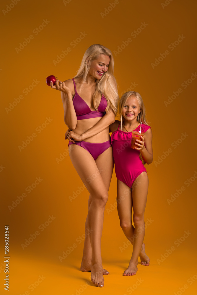 Φωτογραφία blonde woman mom in a swimsuit holds an apple and her daughter in a swimsuit drinks fresh juice
