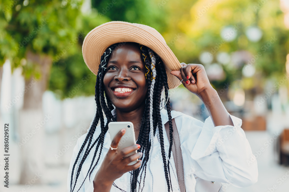 Φωτογραφία Portrait of a smiling young African American girl with pigtails in a hat with a mobile phone walking down the street on a sunny day